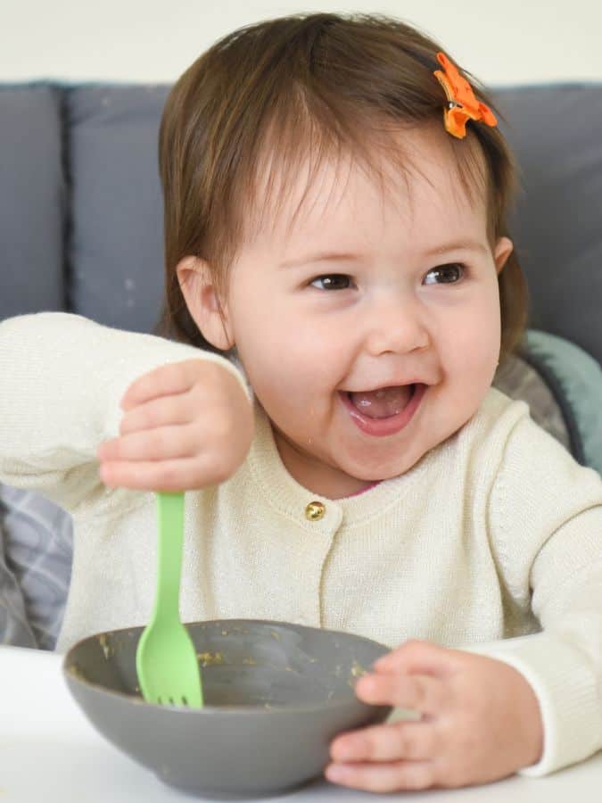 baby happy eating solids from bowl in highchair