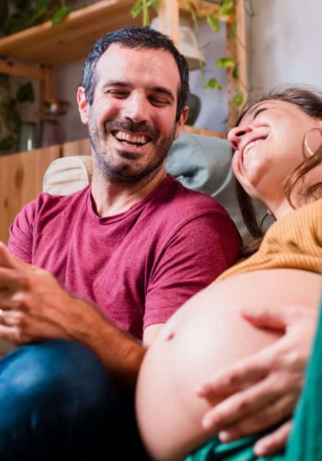 pregnant couple seated on floor leaning into each other and laughing.